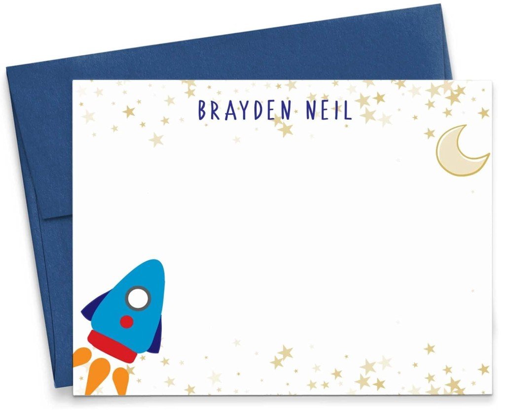 Personalized flat notecard with navy hand-lettered name text, a cartoon rocket ship, gold and yellow stars, a crescent moon, and a matching navy envelope.